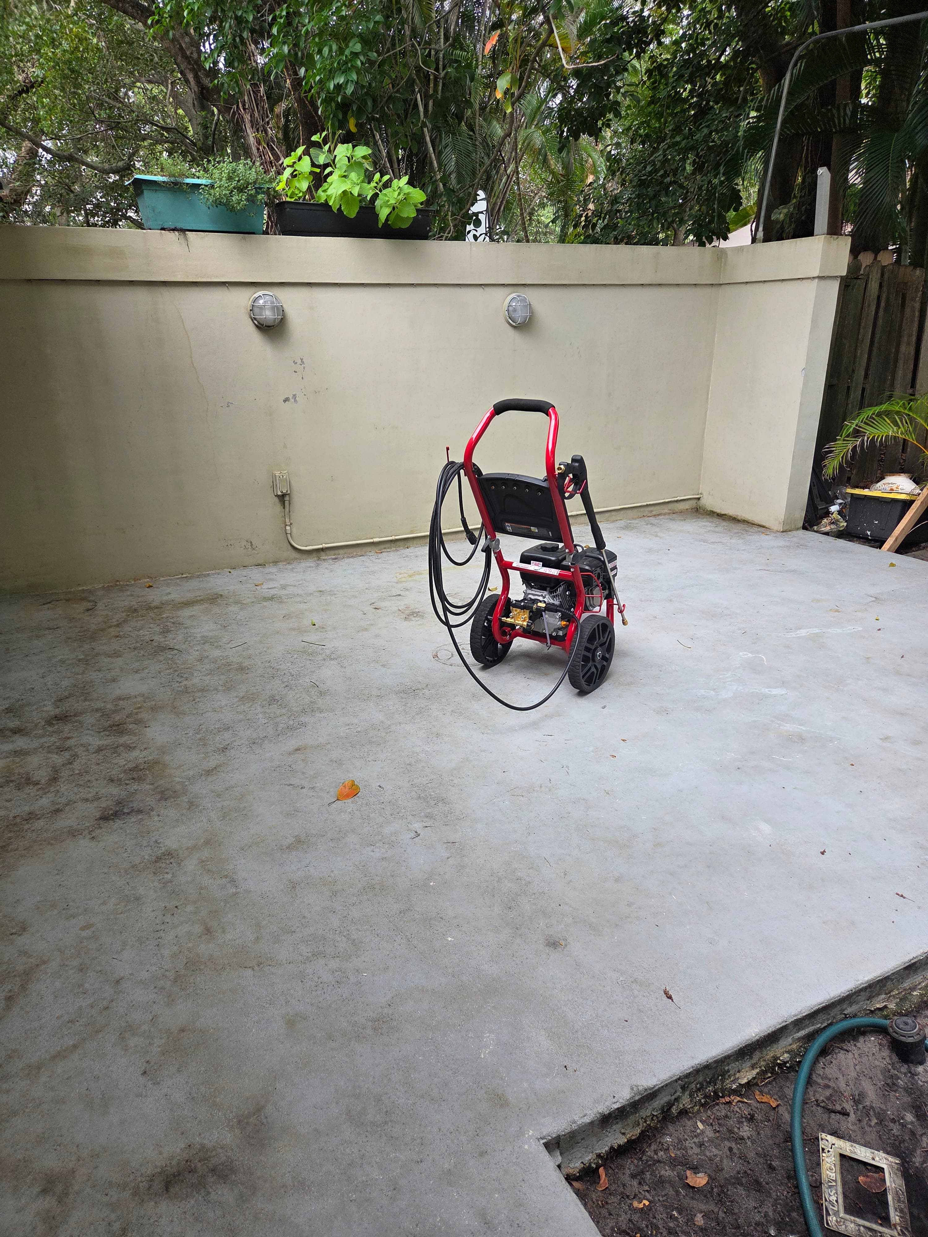 Red pressure washer on a concrete patio with a beige wall and lush green trees.